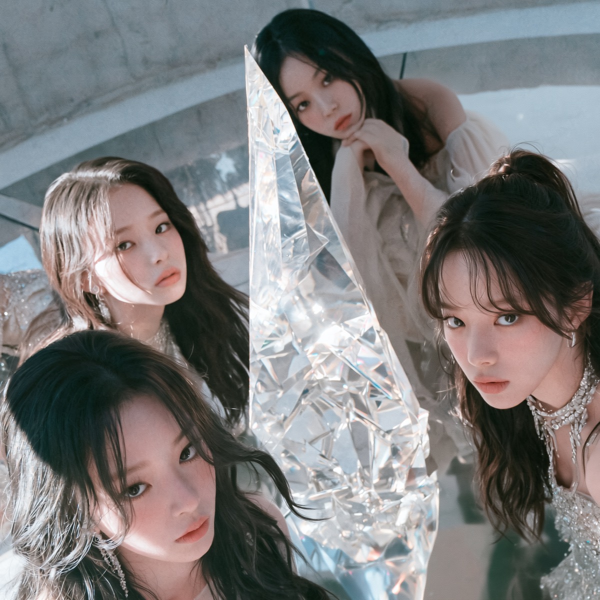 A snapshot of four VIVYD members gathered in a circular room on a pale stone floor, all staring intently into the lens. They lean forward around a tall, jagged prismatic crystal that refracts soft shards of light. One member looks up from the left foreground, two press in on either side in shimmering tops and crystal neckpieces, and a fourth rests her chin on her hands in the softly focused background.